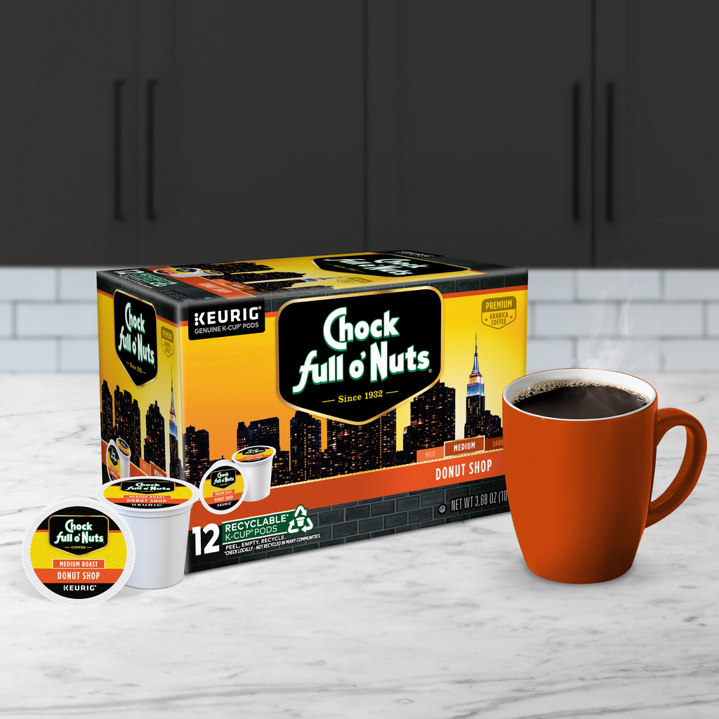 A box of Chock full o'Nuts Donut Shop - Keurig K-Cup® Pods, Medium Roast, rests beside a red mug brimming with steaming coffee on a kitchen countertop.