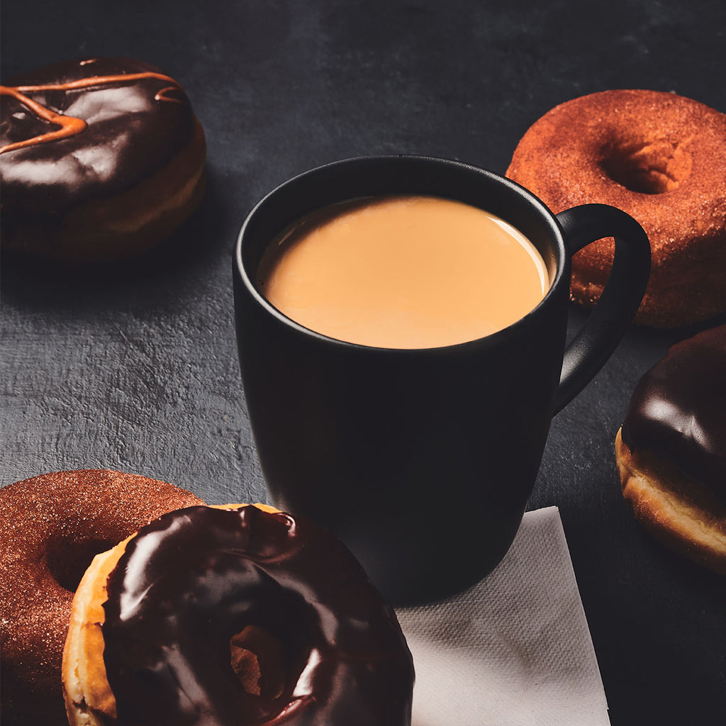 A black mug of 100% Colombian - Medium - Ground coffee by Chock full o'Nuts surrounded by assorted donuts, including chocolate glazed and sugared, placed on a dark surface.