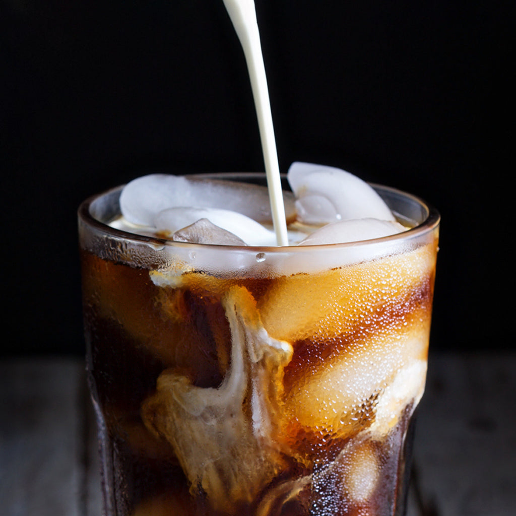 Close-up of a glass filled with iced coffee made from Chock full o'Nuts 100% Colombian - Medium - Ground as milk is being poured in, creating swirling patterns in the dark coffee against a black background.