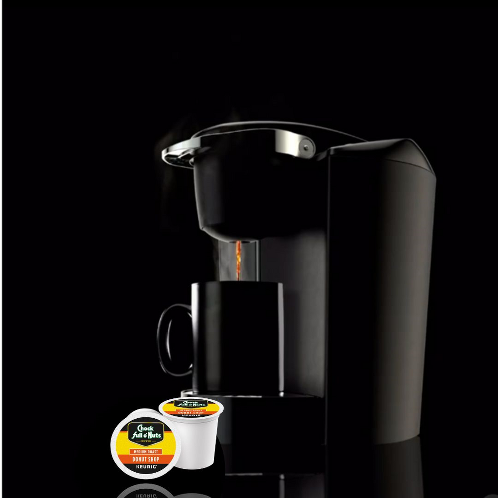 A single-serve coffee maker brews coffee into a mug, with two Chock full o'Nuts Donut Shop - Keurig K-Cup® Pods - Medium Roast placed in front of the machine against a black background.