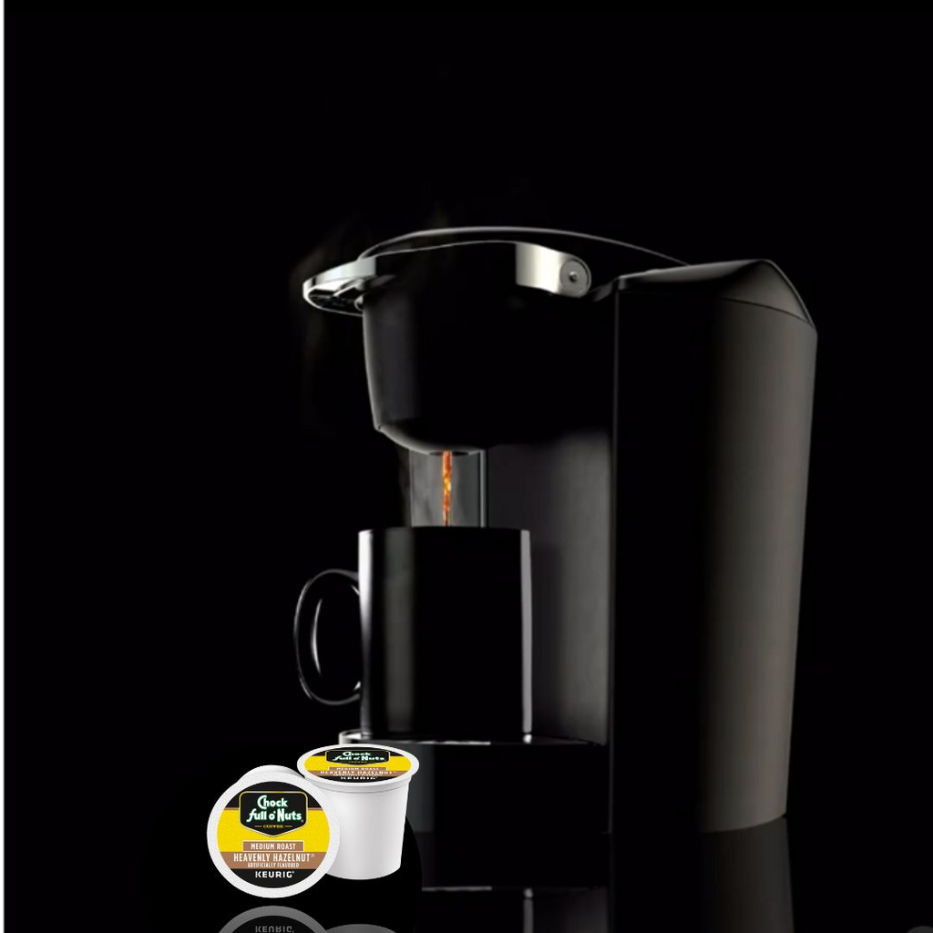 A black single-serve coffee maker brews into a black mug. Two coffee pods labeled "Classic Roast" and "Heavenly Hazelnut - Chock full o'Nuts - Medium Roast" are displayed in front of the machine against a black background.